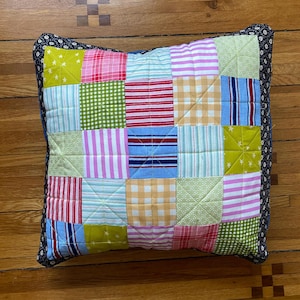 Pode incluir: Uma almofada de patchwork com uma variedade de tecidos coloridos, incluindo rosa, azul, verde, amarelo e vermelho. A almofada &eacute; acolchoada e tem uma guarni&ccedil;&atilde;o floral preta e branca.