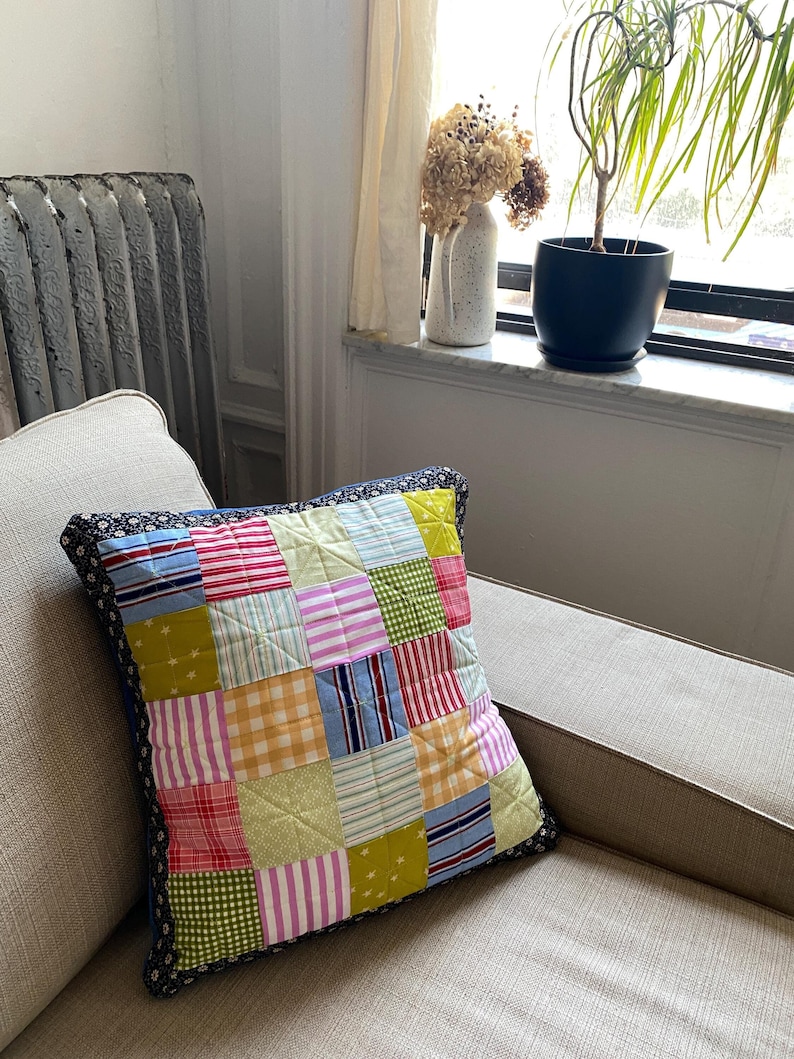 Pode incluir: Uma almofada de patchwork com uma variedade de cores e padr&otilde;es, incluindo vermelho, verde, amarelo, azul e rosa. A almofada tem uma borda azul escura e est&aacute; sentada em um sof&aacute; marrom claro.