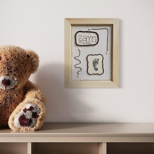 Puede incluir: Un recuerdo enmarcado con la huella del bebé y el nombre. El marco es de madera clara, con un fondo a rayas. El nombre "Zayn" y la fecha "10-13-2025" se muestran en una fuente decorativa. Un oso de peluche marrón está en el fondo.