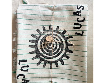 Könnte beinhalten: Gefaltetes weißes Textil mit grünen Streifen, mit einem großen schwarzen Sonnenstrahl-Design und dem Namen "LUCAS". Das Textil ist mit Bindfaden gebunden und hat ein kleines Etikett.