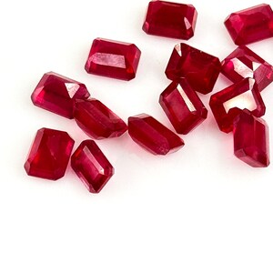 Può includere: Una collezione di rubini sfaccettati, a taglio rettangolare. Le pietre preziose sono di un colore rosso intenso e ricco e sembrano essere sparse su una superficie bianca. I rubini variano leggermente per dimensioni e forma.