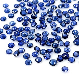 Puede incluir: Un conjunto de pequeñas gemas redondas, de color azul oscuro, dispersas sobre una superficie blanca. Las gemas tienen un aspecto pulido y reflectante, con un color uniforme. La disposición es aleatoria, sin un patrón aparente.