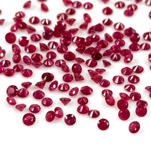 Puede incluir: Una dispersión de pequeñas gemas facetadas de color rojo intenso sobre un fondo blanco. Las gemas son redondas y parecen rubíes, con múltiples facetas que capturan la luz. Las gemas están dispersas.