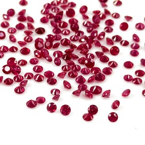 May include: A scattering of small, faceted, deep red gemstones against a white background. The gemstones are round and oval-shaped, with a brilliant cut that catches the light. The gems are scattered randomly, creating a sparkling effect.