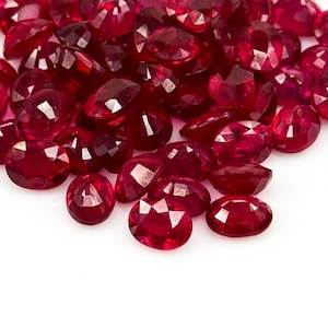 Peut inclure: Un ensemble de rubis ovales et facettés de différentes tailles. Les pierres précieuses sont d'un rouge profond et riche, dispersées sur un fond blanc. Les rubis sont taillés pour maximiser la réflexion de la lumière.