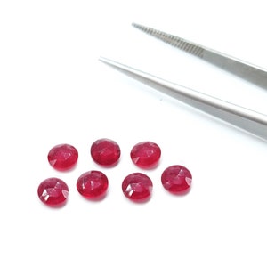 May include: Eight red ruby gemstones, each with a faceted cut, are arranged in a semi-circle on a white surface. A pair of silver tweezers with the word "INOX" printed on them are visible in the background.