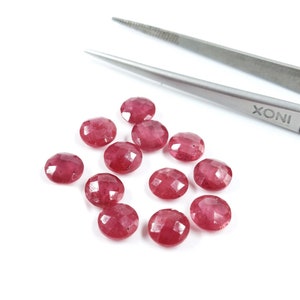 May include: A group of 12 red ruby gemstones, each with a faceted cut, are scattered on a white surface. A pair of silver tweezers with the word "XONI" printed on them are lying on the surface.