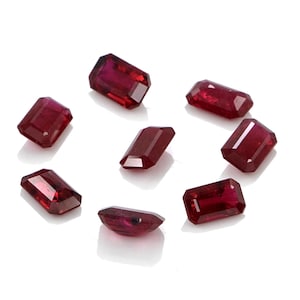 Puede incluir: Un conjunto de siete rubíes sueltos y facetados dispuestos en un patrón circular. Cada piedra preciosa presenta un corte esmeralda rectangular con una coloración roja profunda y superficies reflectantes que captan la luz.