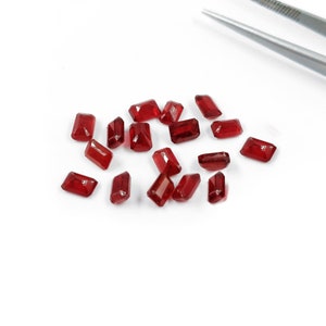 May include: A group of small, rectangular, red gemstones scattered on a white surface. A pair of silver tweezers with the word "INOX" printed on them are visible in the background.