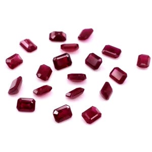 Puede incluir: Un conjunto de rubíes de corte rectangular, dispersos sobre una superficie blanca. Las piedras preciosas muestran un tono rojo intenso, con facetas que capturan la luz. Estos rubíes sueltos son ideales para la creación de joyas.