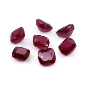 Puede incluir: Una colección de siete rubíes sueltos, tallados en forma de cojín, con un color rojo profundo y saturado. Cada piedra preciosa presenta una superficie facetada que refleja la luz, mostrando una calidad translúcida y un acabado pulido.