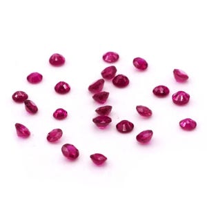 May include: A collection of small, round, faceted, deep red gemstones scattered on a white surface. The gemstones have a polished, reflective surface, and vary slightly in size and shape. The overall effect is one of scattered brilliance.