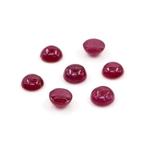 Puede incluir: Seis piedras preciosas pequeñas y redondas con corte cabujón de un color rojo magenta profundo y saturado. Cada piedra presenta una superficie superior abovedada, lisa y pulida, y una base plana, mostrando un aspecto translúcido uniforme.