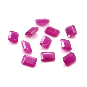 Puede incluir: Una colección de once piedras preciosas sueltas de color rosa vibrante, cada una cortada en forma de esmeralda rectangular con superficies facetadas que reflejan la luz, dispuestas en una formación dispersa sobre un fondo blanco.
