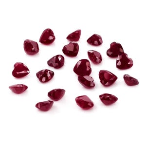 May include: A collection of heart-shaped, faceted rubies scattered on a white surface. The gemstones are a deep, rich red color, with multiple facets catching the light. The shapes vary slightly, adding to their unique appeal.