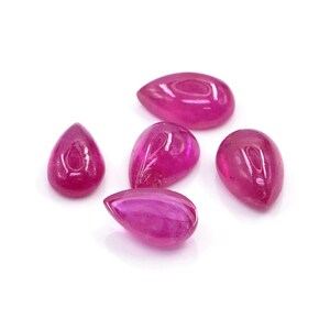 Op de afbeelding: Vijf losse, druppelvormige edelstenen met een levendige, verzadigde magenta-roze tint. Elke steen heeft een glad, gepolijst oppervlak in cabochon-stijl dat licht reflecteert, wat de diepe, uniforme kleur en de afgeronde, symmetrische vorm benadrukt.