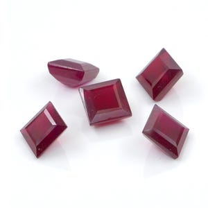 May include: Five loose, faceted gemstones with a deep, saturated red color. Each stone features a distinct geometric shape, including square and elongated diamond cuts, with polished surfaces that reflect light across their angled, flat facets.