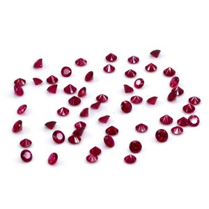 Pode incluir: Uma dispersão de pequenas pedras preciosas vermelhas escuras facetadas sobre um fundo branco. As pedras, de formas e tamanhos variados, apresentam um aspeto polido e brilhante. A imagem evoca luxo e elegância, possivelmente para joias ou elementos decorativos.