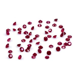 May include: A collection of numerous small, round, faceted loose gemstones in a vibrant, deep red color. Each stone features a brilliant cut with multiple reflective surfaces, scattered across a plain white surface in a loose, unorganized arrangement.