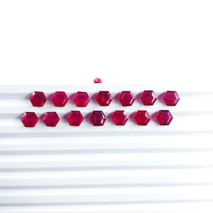 May include: A row of 17 small, faceted, ruby red gemstones arranged in a line on a white surface.