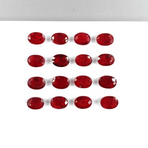 May include: A collection of 15 oval-shaped, red gemstones arranged in three rows of five on a white surface.