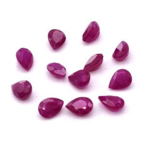 Puede incluir: Un conjunto de gemas de rubí facetadas en forma de pera. Los rubíes son de un color rojo intenso y rico, dispersos sobre una superficie blanca. Las facetas capturan la luz, creando un efecto brillante. Ideal para la fabricación de joyas.
