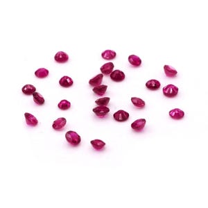 May include: A collection of small, round, faceted, deep red gemstones scattered on a white surface. The gemstones have a polished, reflective surface, and vary slightly in size and shape. The overall effect is one of scattered brilliance.