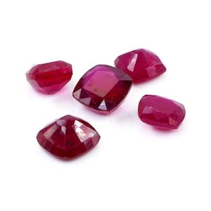 May include: Five loose, cushion-cut rubies with a deep red color and faceted surfaces. Each gemstone displays a polished, reflective finish with sharp edges and a square-like shape, arranged in a cluster against a plain white background.