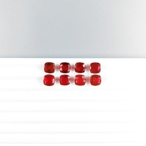 May include: Eight square-cut, vibrant red gemstones are arranged in two rows of four on a white surface. The gems have a polished, reflective surface, and are set against a neutral background.