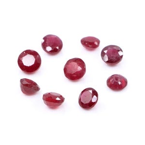 May include: A collection of eight loose, round-cut rubies with a deep red color and faceted surfaces. Each gemstone displays a polished finish and a circular shape, arranged in a scattered pattern against a plain white background.