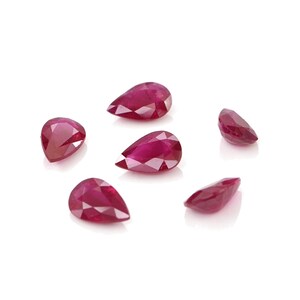 May include: Five loose, pear-shaped rubies with a deep red color and faceted surfaces. The gemstones are arranged in a scattered formation, showcasing their pointed ends and rounded bases, reflecting light across their polished, multi-faceted surfaces.