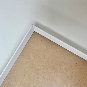May include: Close-up of a corner where a white skirting board meets a light brown floor.