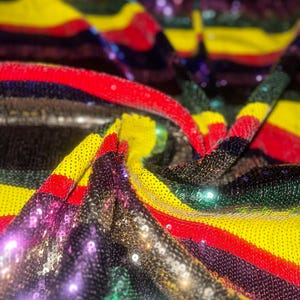 Puede incluir: Primer plano de una tela con lentejuelas de colores. La tela presenta rayas horizontales en negro, rojo, amarillo, verde y morado. Las lentejuelas reflejan la luz, creando un efecto brillante. Ideal para ropa o decoración.