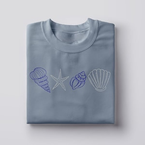 May include: A light blue sweatshirt featuring a line of four seashell illustrations. The designs include a conch shell, starfish, another conch shell, and a scallop shell. The conch shells are dark blue, the starfish is light gray, and the scallop shell is light gray.