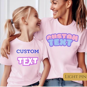 May include: Two light pink t-shirts with the words "CUSTOM TEXT" in a bubble font. The text on one shirt is blue and purple, and the other is blue and pink. The shirts are worn by two people.