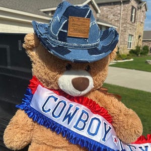 May include: A brown teddy bear wearing a denim cowboy hat with a Levi Strauss & Co. patch. The bear has a red, white, and blue sash that says "COWBOY". The bear is sitting outside.