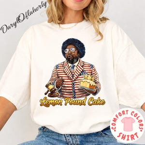 Puede incluir: Camiseta Comfort Colors color crema con una ilustración de dibujos animados de un hombre con un traje rojo, blanco y azul sosteniendo un mazo y un pastel con una corona. El texto "Lemon Pound Cake" está debajo de la ilustración. Un logotipo de camiseta Comfort Colors rosa está en la parte inferior derecha.