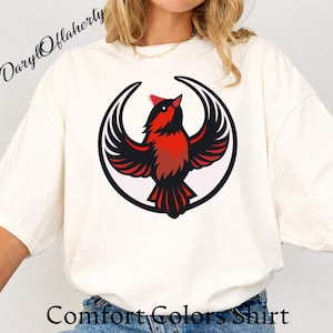 May include: Cream-colored Comfort Colors shirt featuring a red and black bird design within a black circle. The bird has outstretched wings. The text "Comfort Colors Shirt" is at the bottom of the shirt. The name "Daryl Oflaherty" is at the top left.