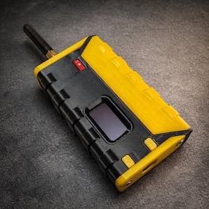 Puede incluir: Un dispositivo electrónico negro y amarillo con una pequeña pantalla y una antena. El dispositivo tiene forma rectangular con una parte superior y laterales amarillos texturizados, y una parte delantera y trasera negras. Tiene un interruptor "ON".