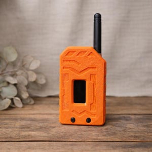 May include: An orange walkie-talkie with a black antenna and two black buttons. The walkie-talkie has a textured surface and a rectangular screen. The device is standing on a wooden surface.
