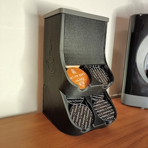 May include: A black coffee pod holder with two dispensing slots. The holder has a textured surface and a gray top. The holder is filled with coffee pods, including one labeled "CAFÉ AU LAIT". A coffee cup design is on the side.