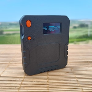 Op de afbeelding: Een zwart, vierkant elektronisch apparaat met een getextureerde afwerking. Het is voorzien van twee oranje ronde knoppen aan de linkerkant en een klein digitaal scherm met tekst waaronder 'heap', 'Flash', 'Ver. 2.7.28' en 'Up 5m' in het blauw.