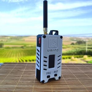 Könnte beinhalten: Ein tragbares elektronisches Gerät mit einem weißen und schwarzen rechteckigen Gehäuse, einer vertikalen schwarzen Antenne, einem kleinen Bildschirm und dem aufgedruckten Text VIRIATO, PRG und RST. Das Gerät verfügt über seitliche Lüftungsschlitze.