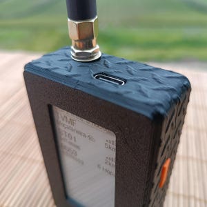 Puede incluir: Dispositivo electrónico negro con superficie texturizada y pantalla pequeña con texto. Una antena está unida a la parte superior. El dispositivo tiene un botón naranja en el lateral y un puerto USB-C.