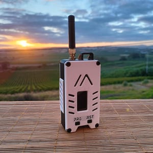 May include: A rectangular electronic device with a white front panel and black sides, featuring a vertical black antenna, a small screen, and two buttons labelled PRG and RST. The front panel has a triangular logo and side ventilation slots.