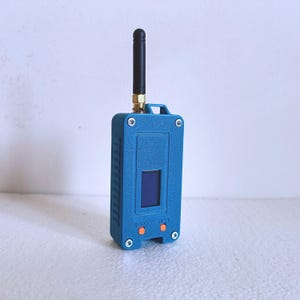 Peut inclure: Un appareil électronique bleu rectangulaire doté d'une antenne noire sur le dessus, d'un petit écran sombre et de deux boutons orange étiquetés PRG et RST. Le boîtier est fixé par quatre vis argentées aux coins et possède une attache supérieure.