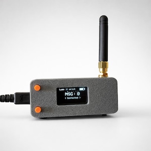 以下が含まれることがあります： 「MSG: 0」と「< Connected >」を表示する小さな画面を備えたグレーの電子デバイス。オレンジ色のボタンが2つ、黒いアンテナ、USBケーブルがあります。「Luso 3D print」の文字も画面に表示されています。