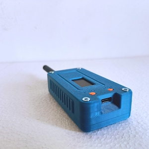 Op de afbeelding: Een rechthoekig blauw elektronisch apparaat met een getextureerde afwerking, voorzien van een klein zwart scherm, twee oranje knoppen met de labels PRG en RST, een zwarte antenne en een USB-poort. De behuizing is vastgezet met vier zilveren schroeven.