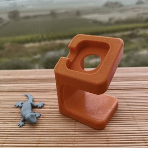 Könnte beinhalten: Ein orangefarbener 3D-gedruckter Ständer für eine Smartwatch. Der Ständer hat eine abgerundete quadratische Form mit einem kreisförmigen Ausschnitt für das Zifferblatt der Uhr. Der Ständer steht auf einer braunen Bambusmatte.