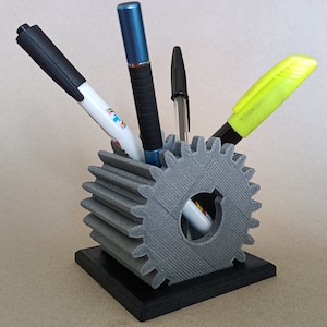 May include: A gray 3D printed gear-shaped pen holder with a black base. The holder has four slots for pens and pencils. There are four writing utensils in the holder: a black marker, a blue pen, a black pen, and a yellow highlighter.